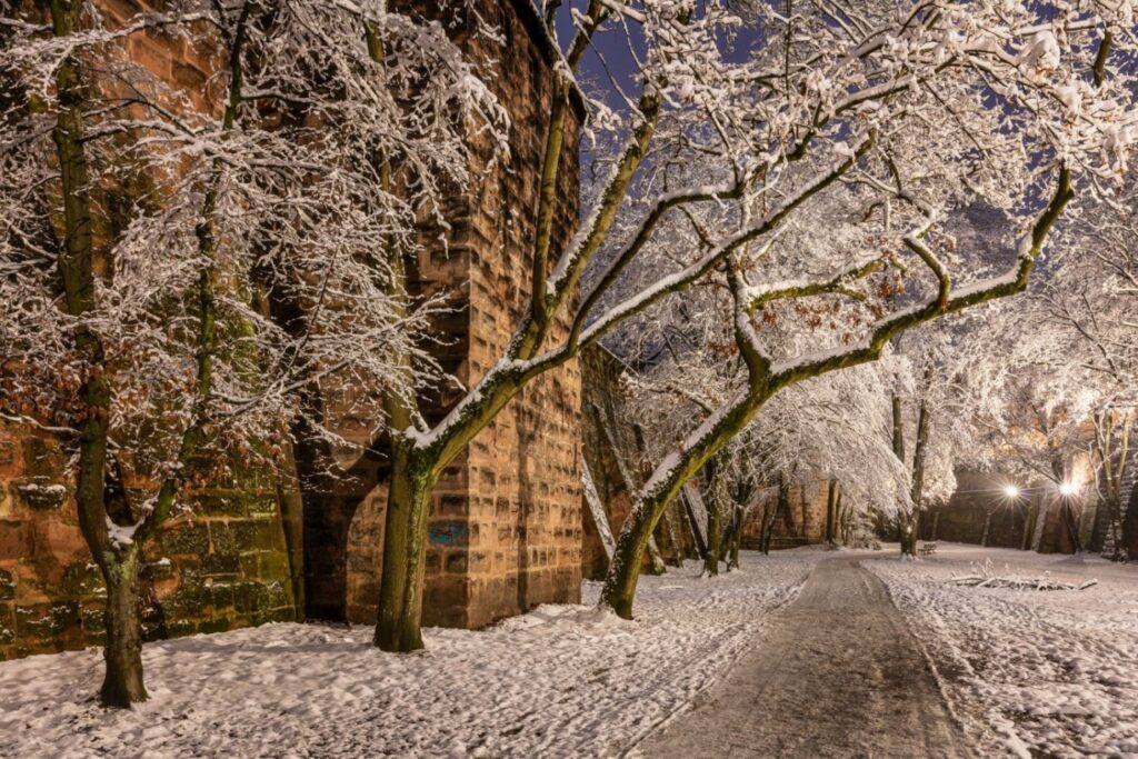 Ein schneebedeckter Weg führt nachts entlang einer alten Steinmauer, umgeben von stark verschneiten Bäumen, die im warmen Licht der Laternen erstrahlen.
