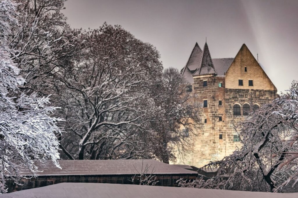 Das Bild zeigt eine hell erleuchtete alte Burg, die von zahlreichen schneebedeckten Bäumen in einer Winternacht umgeben ist.