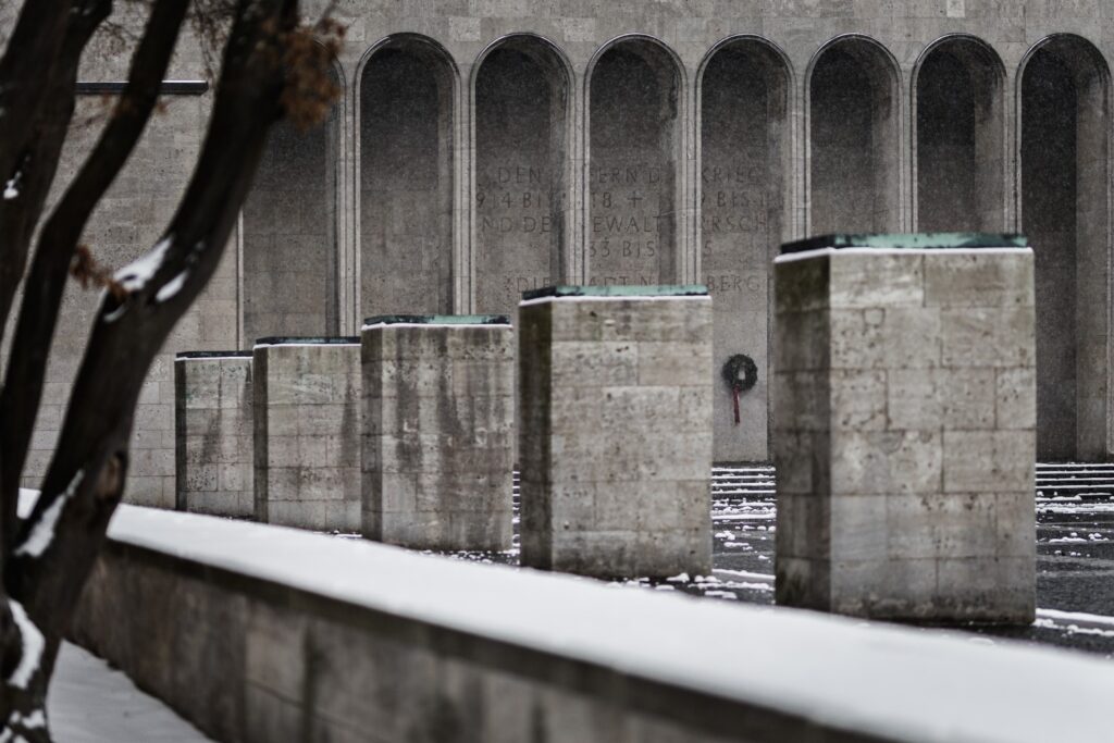 Schneebedeckte Gedenkstätte: Reihe grauer Betonpfeiler vor Wand mit bogenförmigen Nischen.