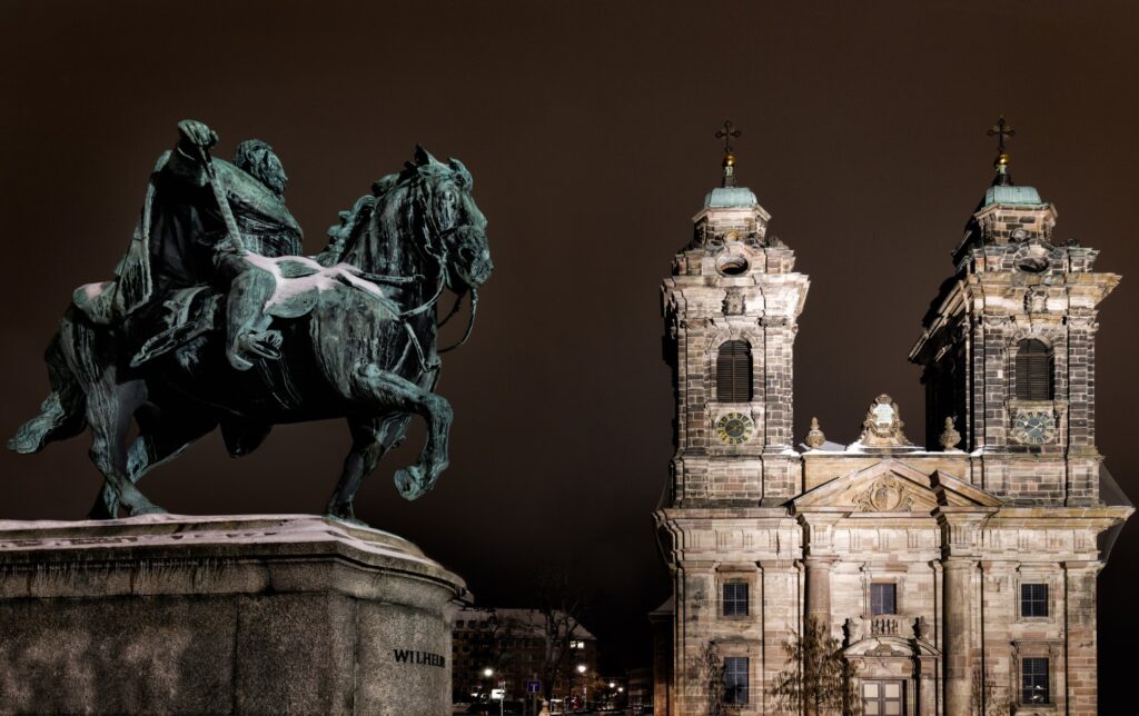Nachtaufnahme einer bronzenen Reiterstatue neben einer barocken Kirche mit zwei Türmen.