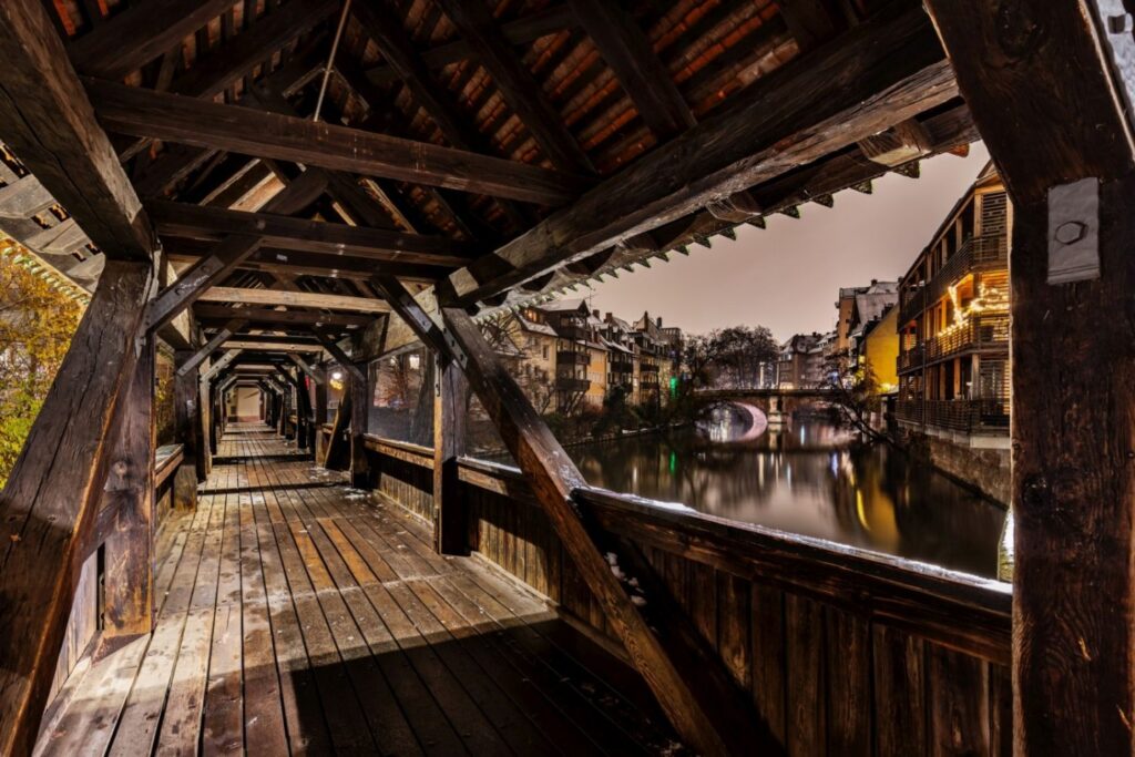 Das Bild zeigt den Blick aus dem Inneren einer langen, überdachten Holzbrücke auf einen Fluss, an dessen Ufern bei Dämmerung oder Nacht beleuchtete Gebäude und eine weitere Bogenbrücke zu erkennen sind.