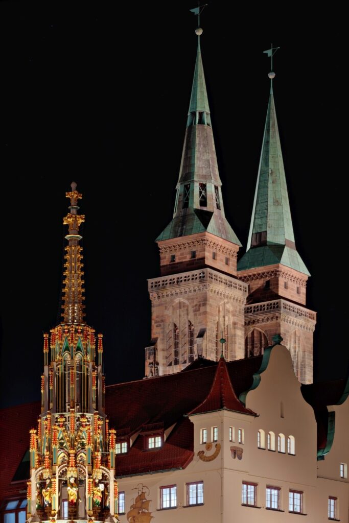 Nachtaufnahme der markanten Türme der Nürnberger Kirche mit kunstvollem, beleuchtetem Denkmal im Vordergrund, das die historische Architektur der Stadt widerspiegelt.