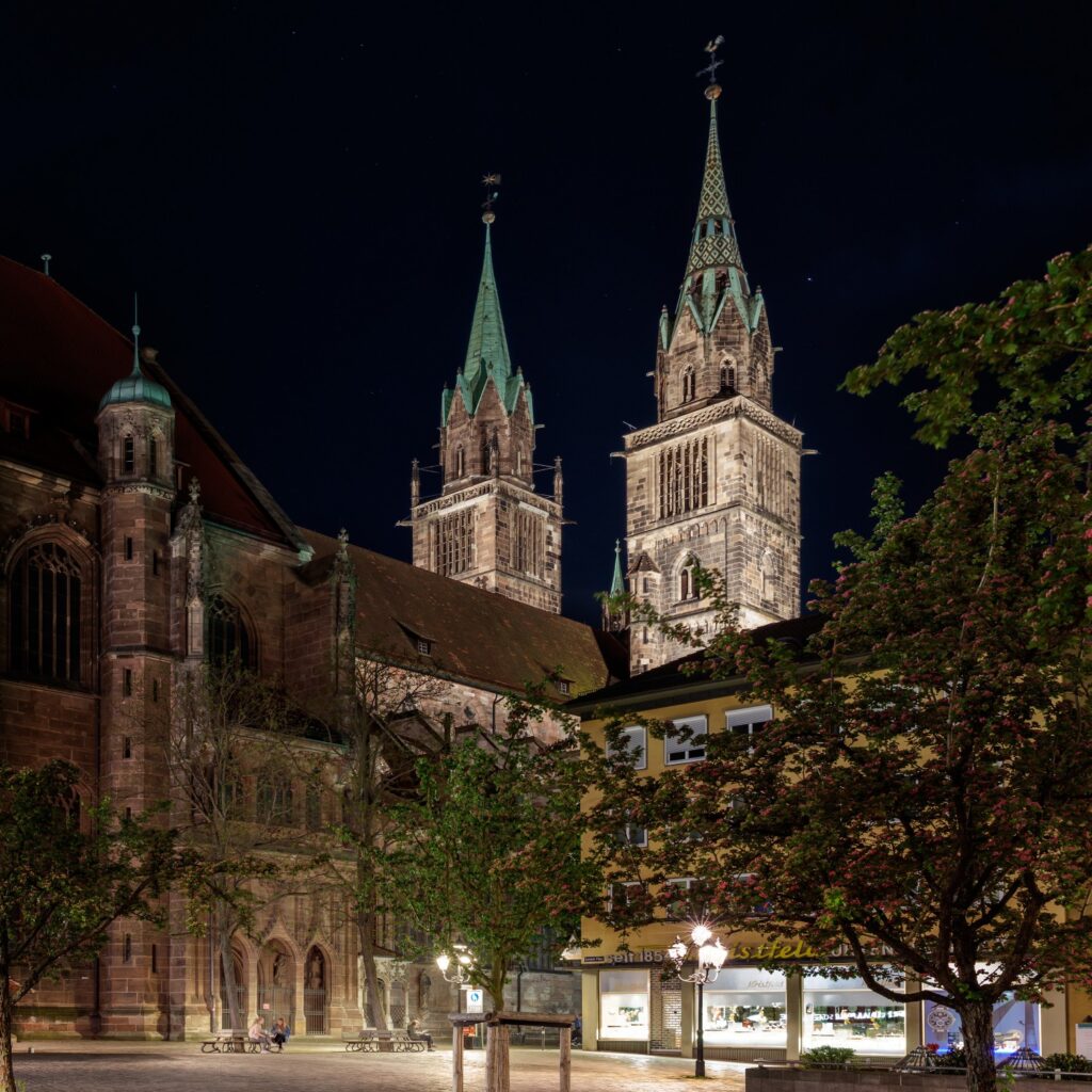 Nachtaufnahme einer gotischen Kathedrale mit zwei Türmen und kupfergrünen Dächern, beleuchtete Stadtszene mit Bäumen.