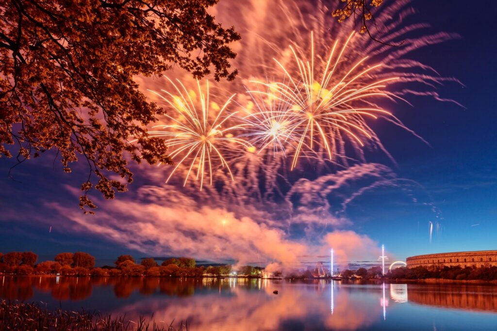 Feuerwerk über einem ruhigen See bei Sonnenuntergang, umgeben von Bäumen und reflektierenden Lichtern, mit dramatischem Himmel und Wolkenformationen.
