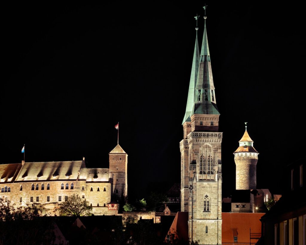 Nachtaufnahme einer historischen Stadt mit gotischem Kirchturm und beleuchteter Festung im Stadtzentrum.