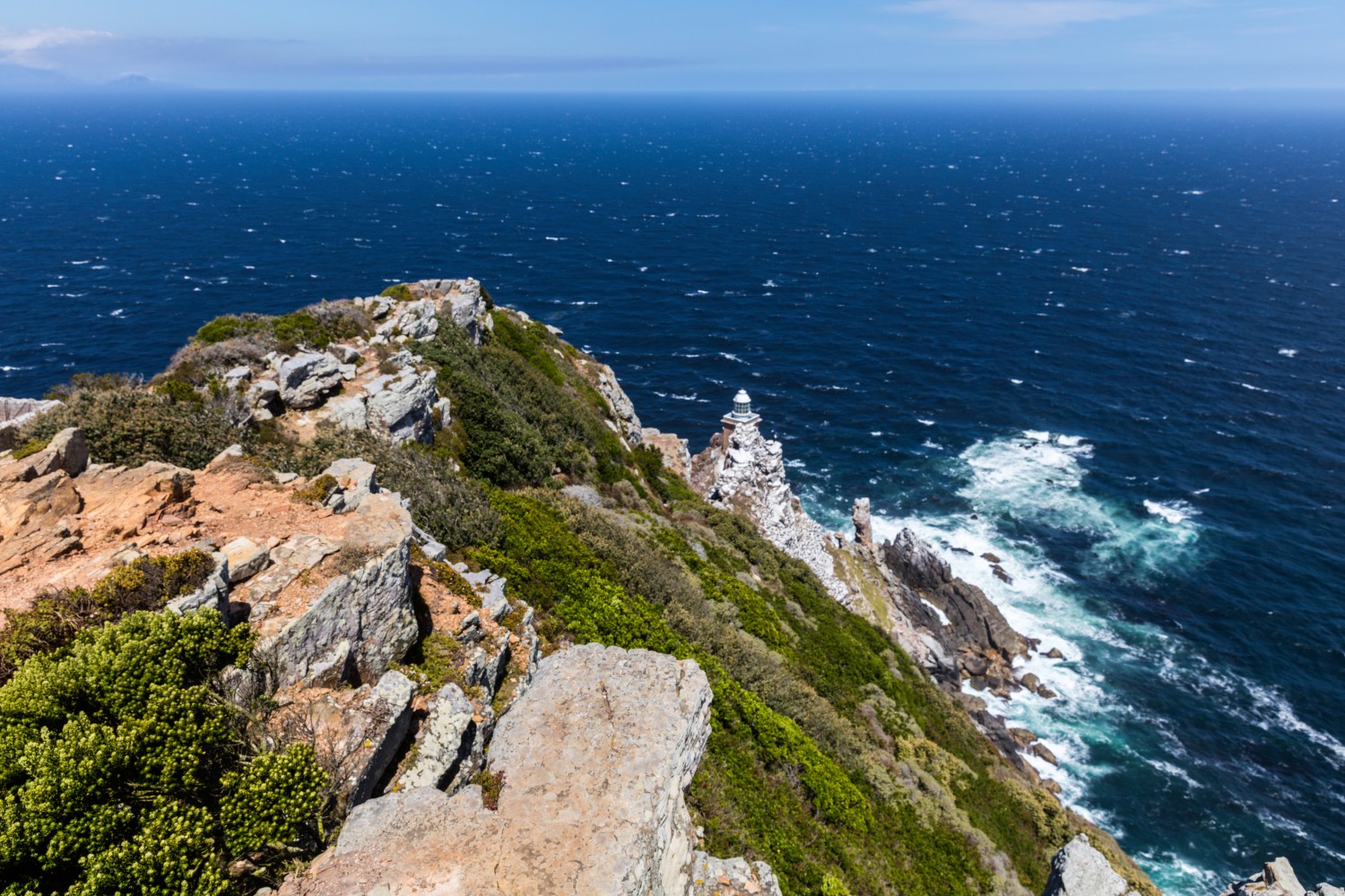Klippenküste mit kleinem Leuchtturm, Felsen und grünem Bewuchs, Aussicht aufs weite blaue Meer.