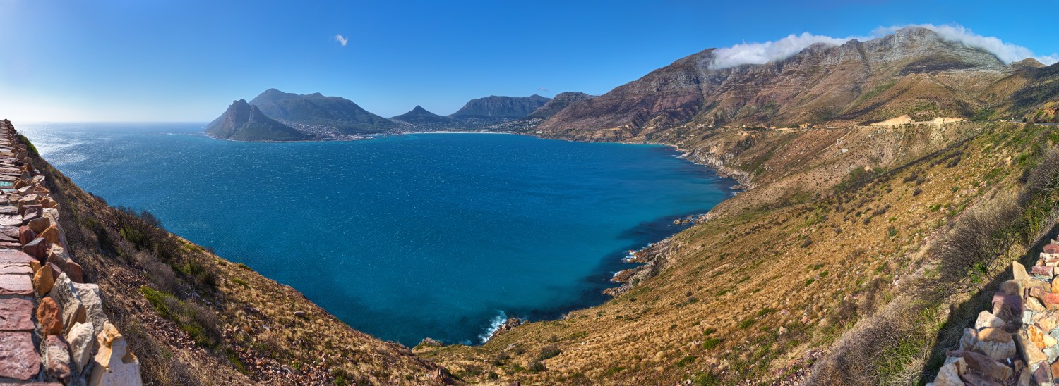 Küstenpanorama mit blauem Meer, felsigen Bergen und steilen Klippen; linke Steinmauer säumt den Pfad.