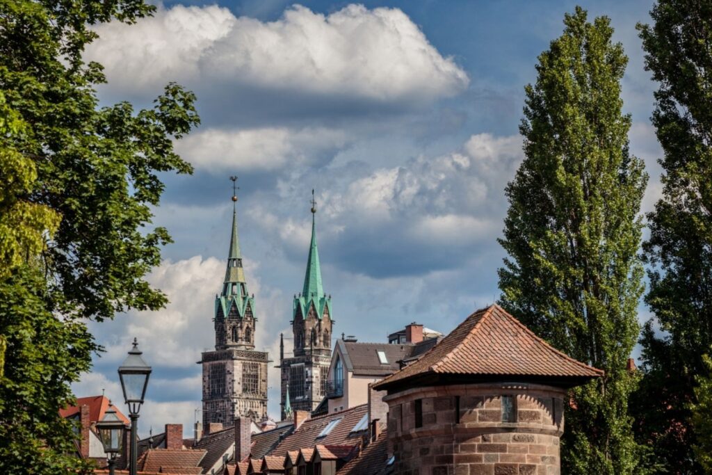 Stadtansicht von Nürnberg mit markanten Türmen und historischen Gebäuden, umgeben von Bäumen und blauem Himmel mit Wolken.
