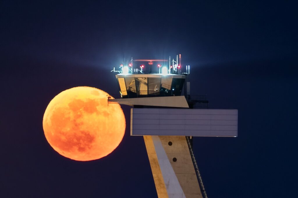 Tower eines Flughafens mit einem leuchtenden, orangen Vollmond im Hintergrund, bei Nacht.