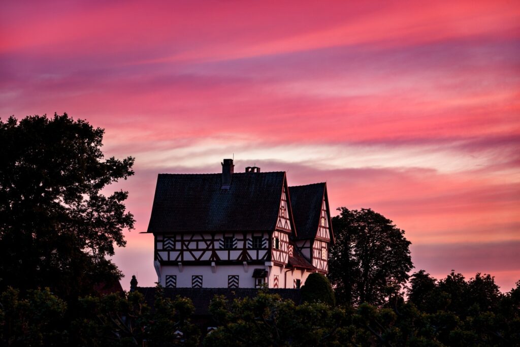Fachwerkhaus vor einem farbenprächtigen Sonnenuntergang mit pinken und lila Wolken, umgeben von Bäumen und Grünanlagen.