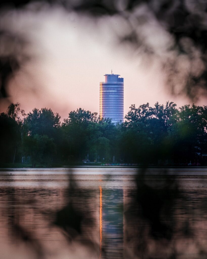 Ein hoher, zylindrischer Glasturm ragt bei Sonnenuntergang hinter einer Baumreihe empor. Im Vordergrund liegt ein ruhiger See, in dem sich das Gebäude als schmaler, orangefarbener Lichtstreifen spiegelt. Unscharfe dunkle Blätter rahmen das Bild oben und unten ein und verleihen der Szene eine natürliche, leicht verträumte Atmosphäre.