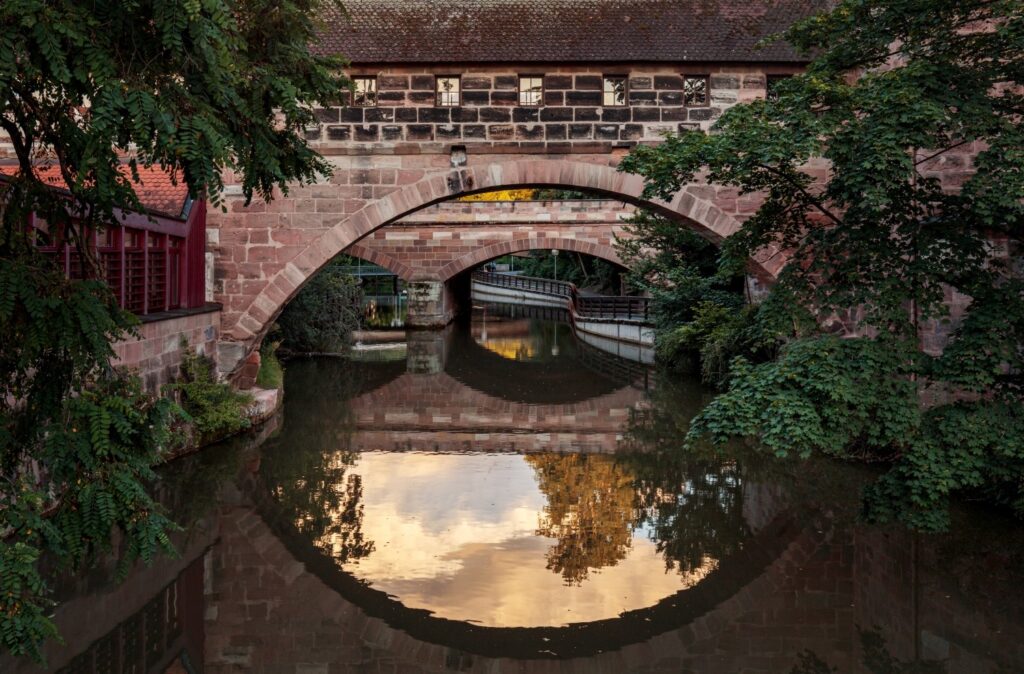 Über einem ruhigen Fluss erstreckt sich eine steinerne Brücke mit mehreren Bögen, die Teil eines Gebäudes mit Fenstern ist und von grünen Bäumen umrahmt wird, wobei sich alles im glatten Wasser golden spiegelt.