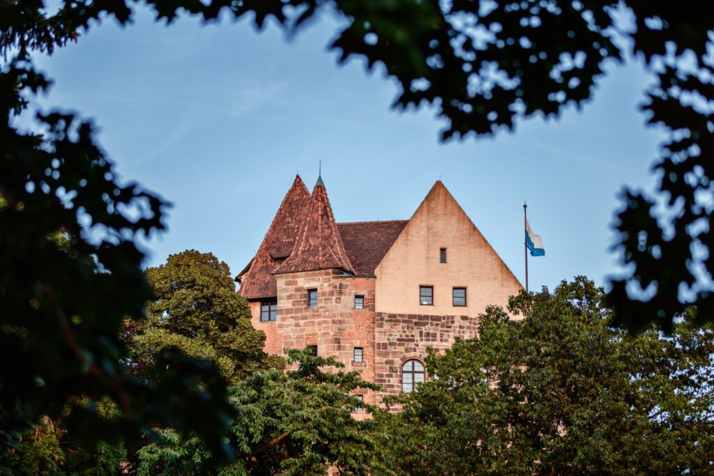 Historisches Schloss aus rotem Sandstein, Türme, von Bäumen umgeben, blauer Himmel und Flagge im Vordergrund.