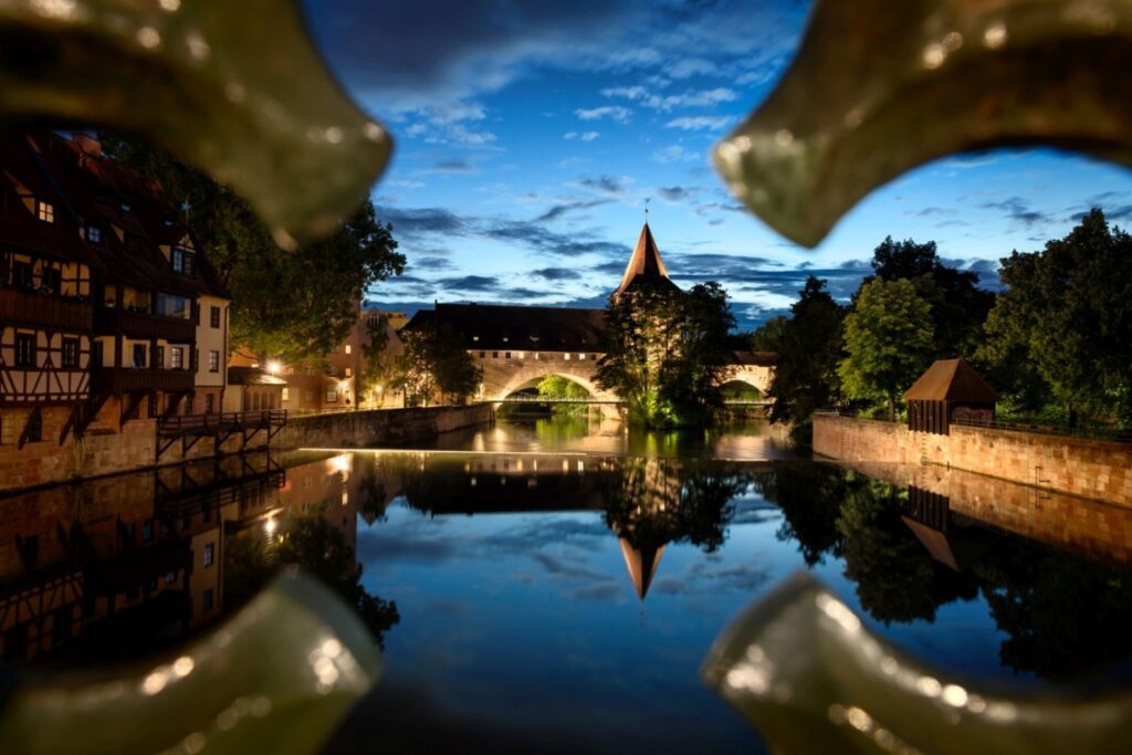 Gerahmt von unscharfen Elementen im Vordergrund zeigt das Bild eine romantische Flusslandschaft bei Dämmerung mit einer beleuchteten Bogenbrücke, einem markanten Turm und malerischen Häusern, die sich im Wasser widerspiegeln.