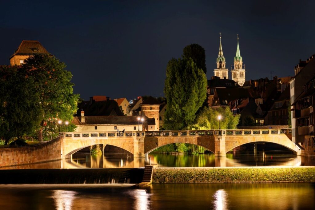 Eine nächtliche Stadtansicht zeigt eine beleuchtete Steinbrücke über einem Fluss, umgeben von Gebäuden und Bäumen, mit zwei markanten Kirchtürmen am Horizont.