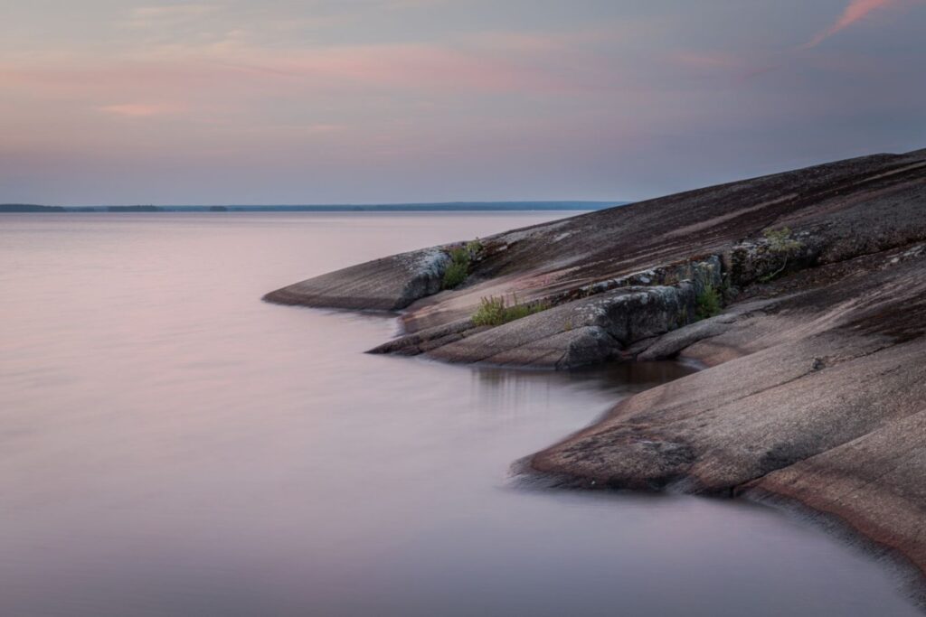 Sanftes Seeufer mit glatten Felsen, ruhiges Wasser und Pastellhimmel beim Sonnenuntergang – Landschaftsfoto am See.