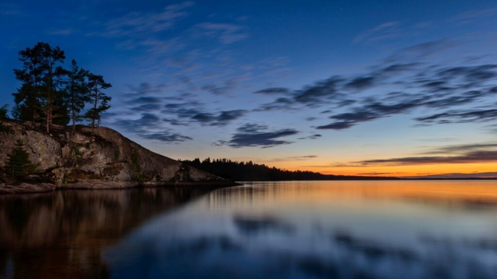 Abendlicher See mit felsigem Ufer, Kiefernwald und ruhiger Spiegelung des Himmels im Sonnenuntergang.