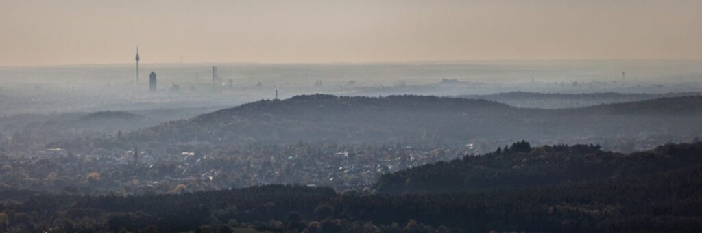 Panoramablick über bewaldete Hügel, Dorf im Vordergrund, entfernte Stadt-Skyline am nebligen Horizont.