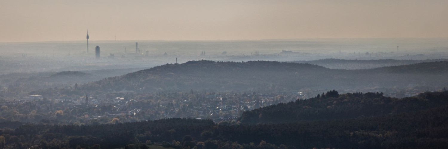 Panoramablick über bewaldete Hügel, Dorf im Vordergrund, entfernte Stadt-Skyline am nebligen Horizont.