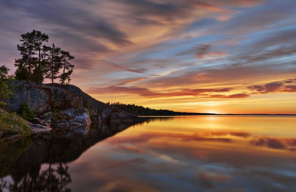 Sonnenuntergang über einem ruhigen See mit reflektierenden Farben und silhouettierten Bäumen am Ufer.
