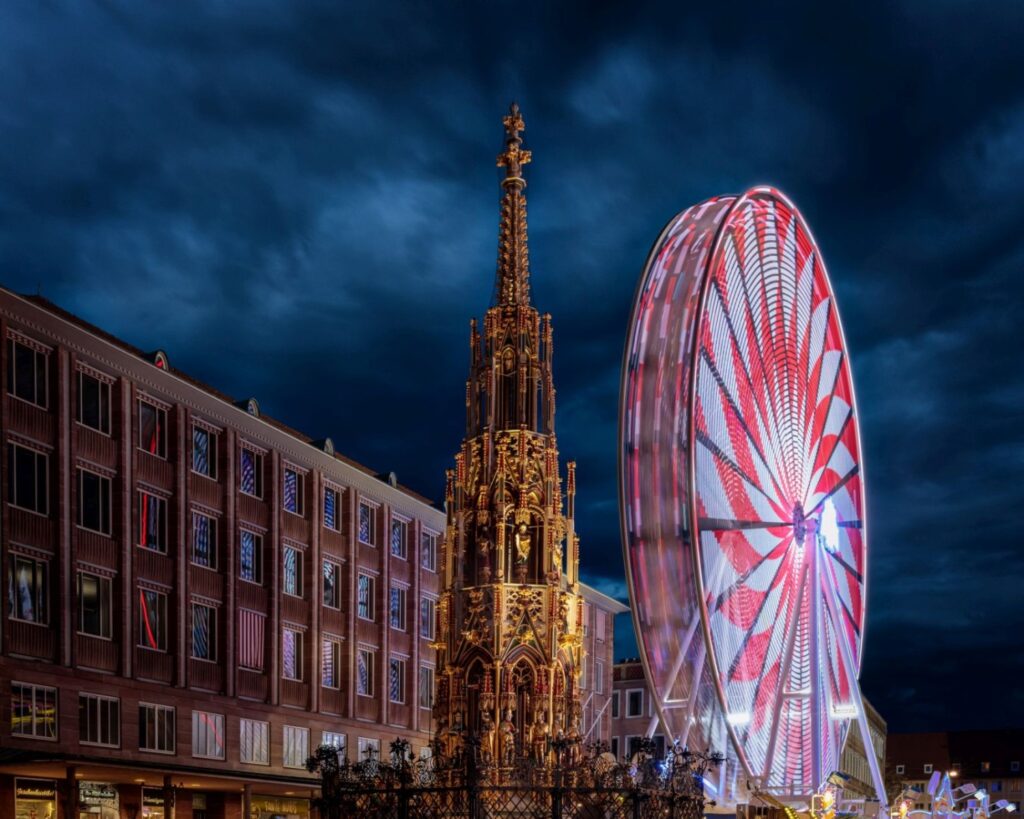 Die Nachtaufnahme zeigt einen reich verzierten Brunnen und ein leuchtendes, sich drehendes Riesenrad vor einem dunkelblauen Wolkenhimmel.