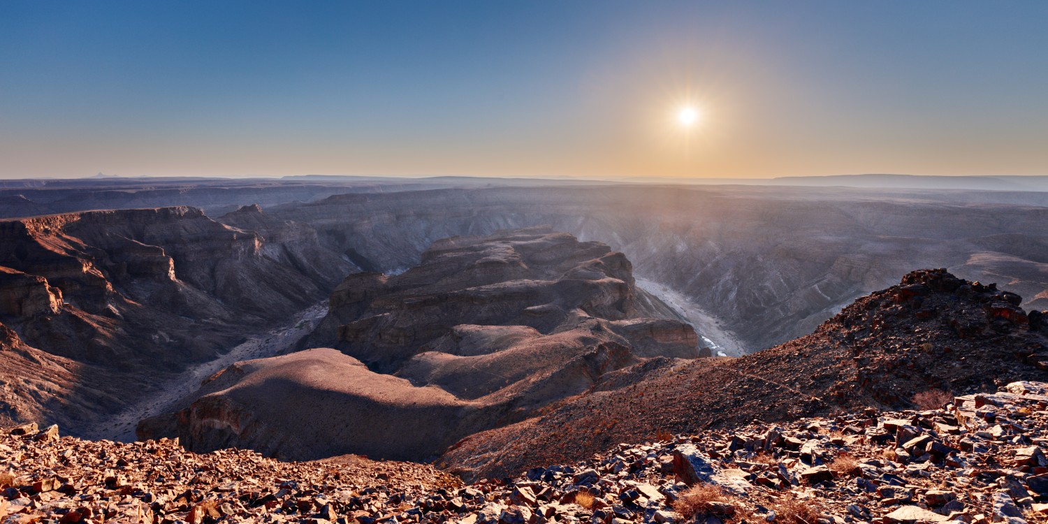 Weite Wüsten-Canyonlandschaft bei Sonnenaufgang, rote Felsformationen, tiefe Schluchten, trockenes Flussbett.