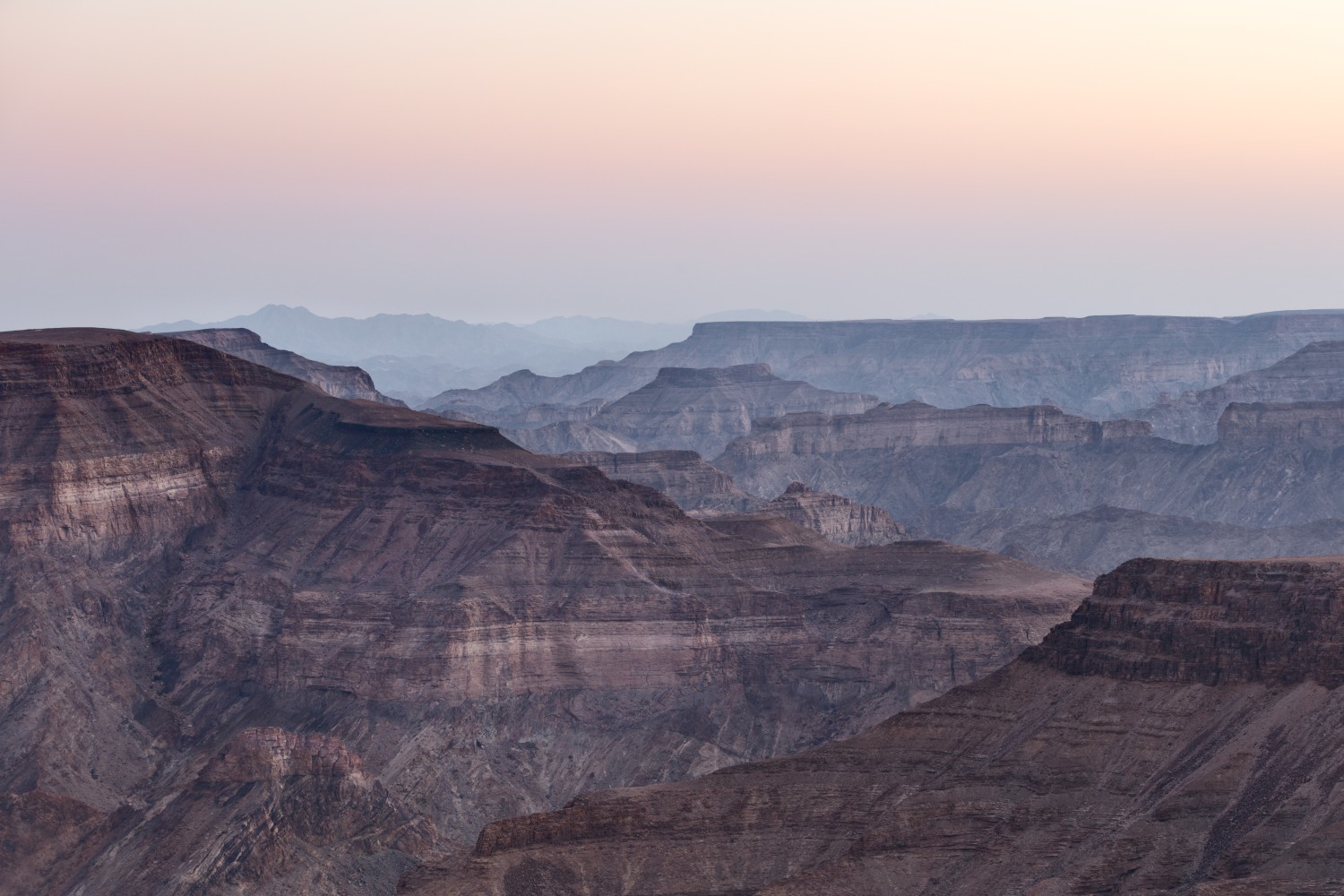 Weite Canyonlandschaft mit schichtigen Felswänden und sanftem Sonnenaufgangshimmel in Pastellfarben.