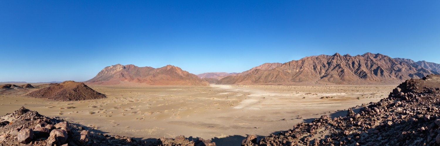 Panorama einer Wüstenlandschaft mit sandiger Ebene, Felsbrocken und Bergkette unter klarem blauen Himmel.