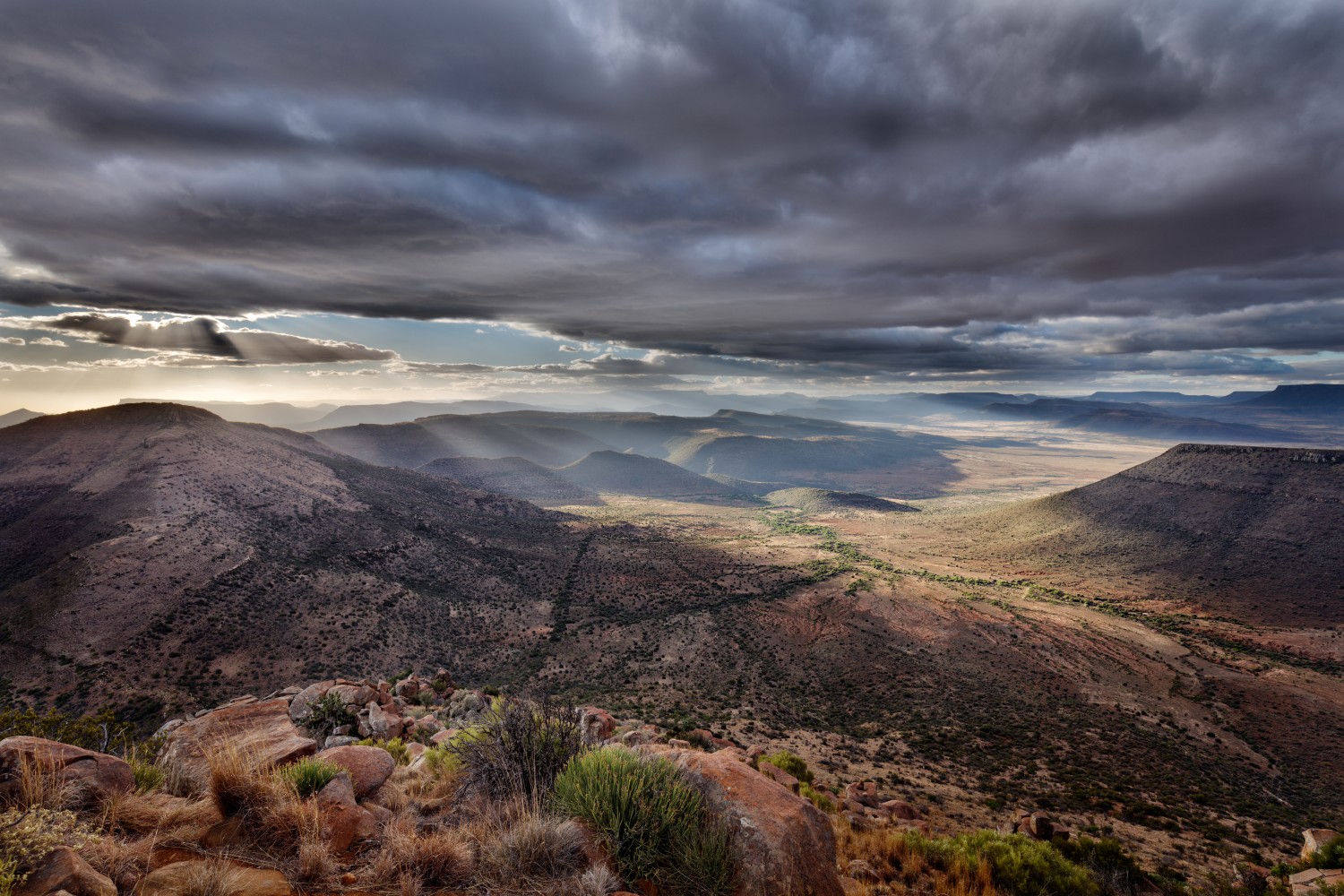 Panorama einer Wüstenlandschaft mit Hügeln, Felsen, grünem Tal und Sonnenstrahlen durch dunkle Wolken.