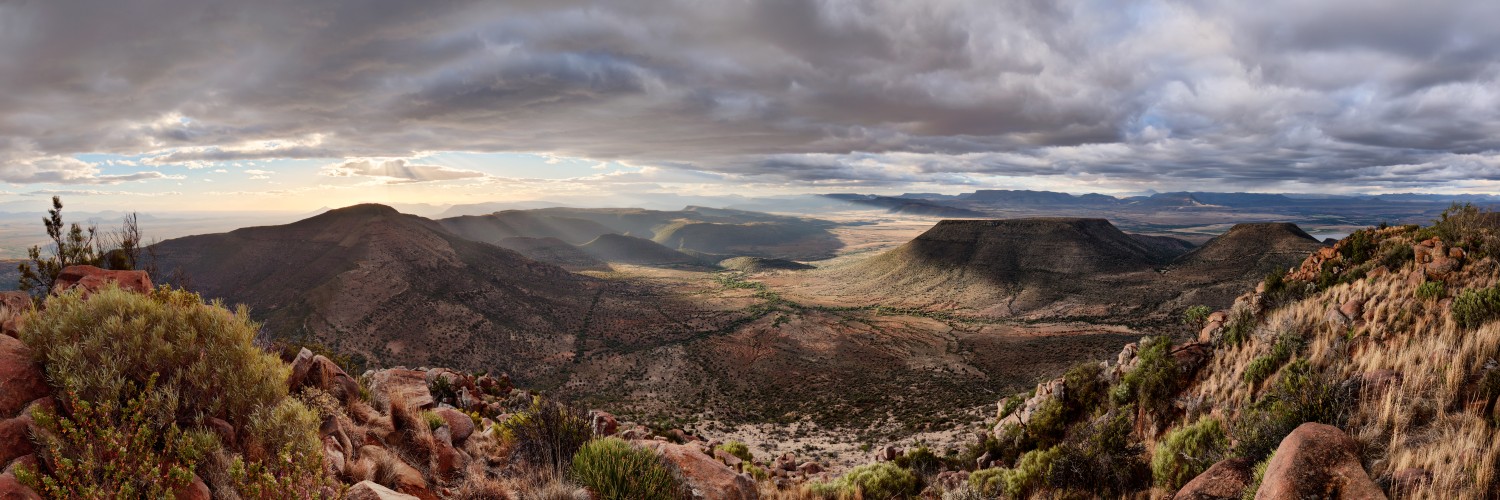 Panorama einer Wüstenlandschaft mit Mesa-Berge, Felsbrocken, Buschwerk und dramatischem Himmel über dem Tal.