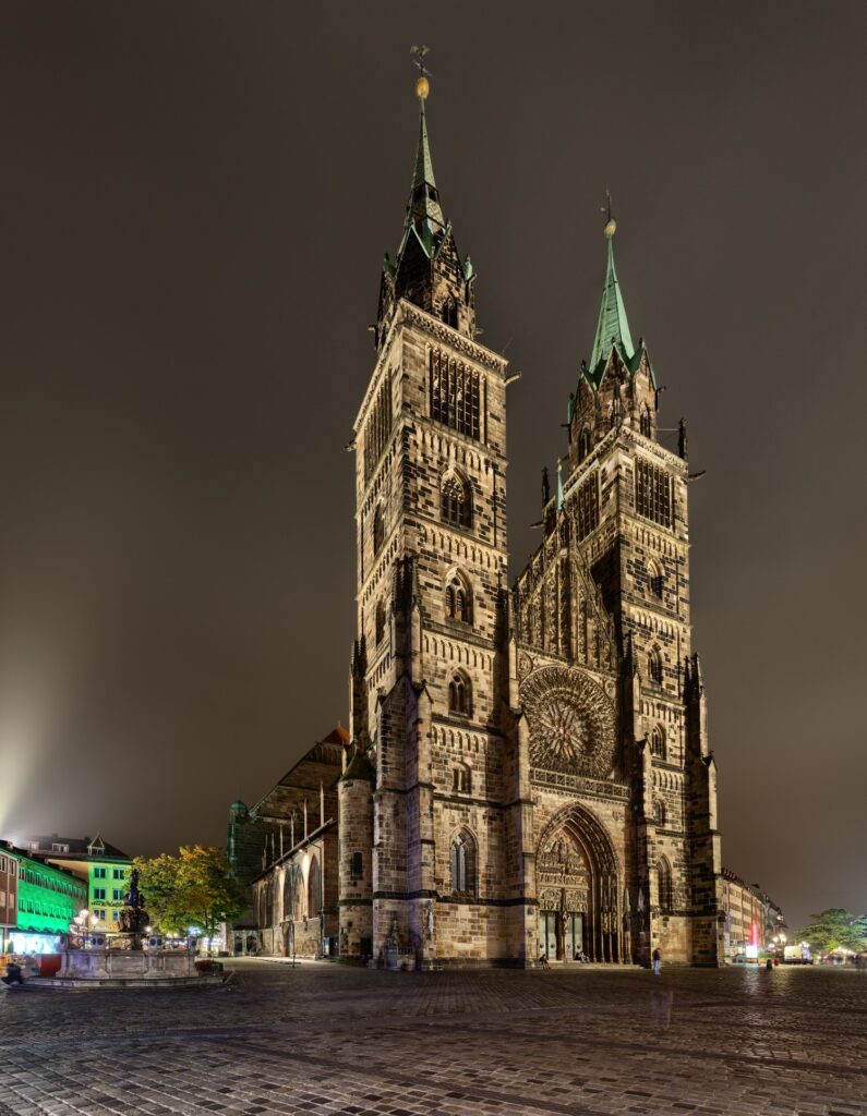 Nachtaufnahme einer gotischen Kathedrale mit zwei Türmen, Rosettenfenster und beleuchteter Steinfassade auf dem Platz.