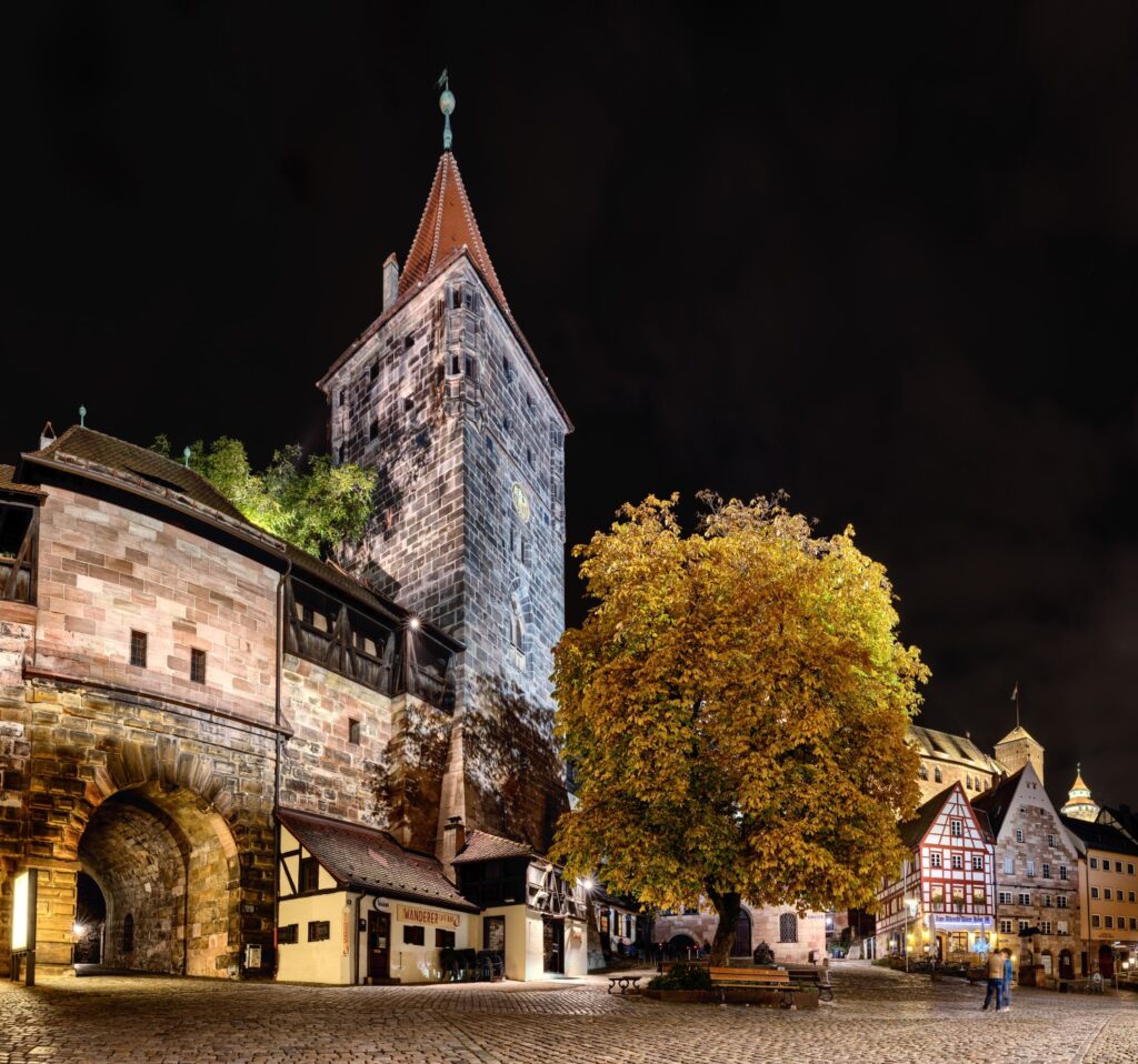 Nachtaufnahme einer historischen Stadt: gotischer Torturm mit rotem Dach, Fachwerkhäusern und Herbstbaum am Kopfsteinpflaster.