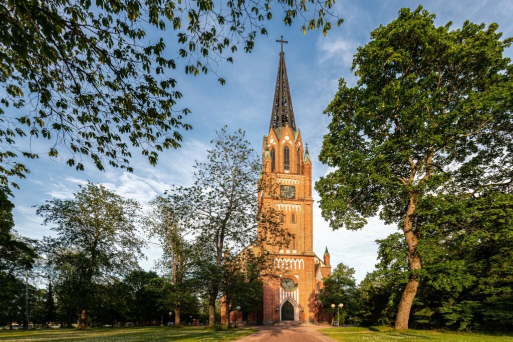 Eine markante Backsteinkirche mit einem hohen spitzen Turm und einer Uhr dominiert das Zentrum des Bildes, umgeben von einer grünen Parklandschaft mit Bäumen unter blauem Himmel, wobei grüne Äste das obere Bildfeld rahmen und ein Weg zum Eingang führt.