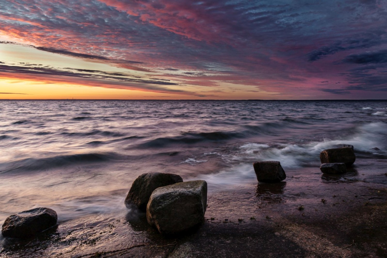 Sonnenuntergang über ruhigem Meer mit Felsen am Ufer, farbiger Himmel in Orange, Lila und Blau.