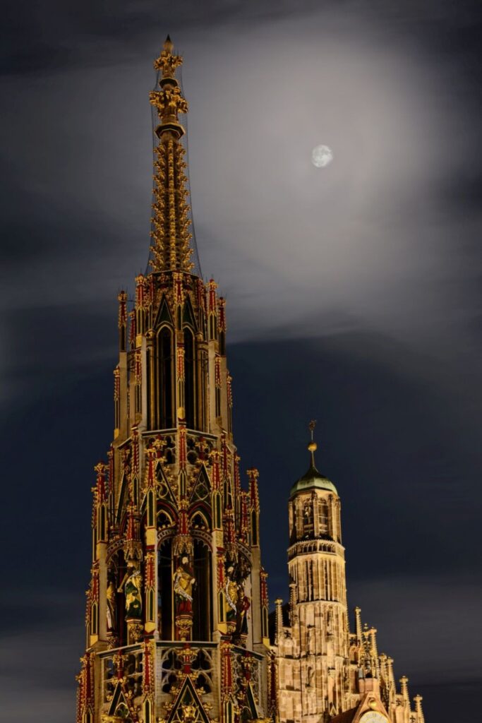 Ein prächtig goldener und verzierter Turm ragt neben einem steinernen Turm mit grüner Kuppel unter einem von Wolken bedeckten Himmel mit einem sichtbaren Mond empor.