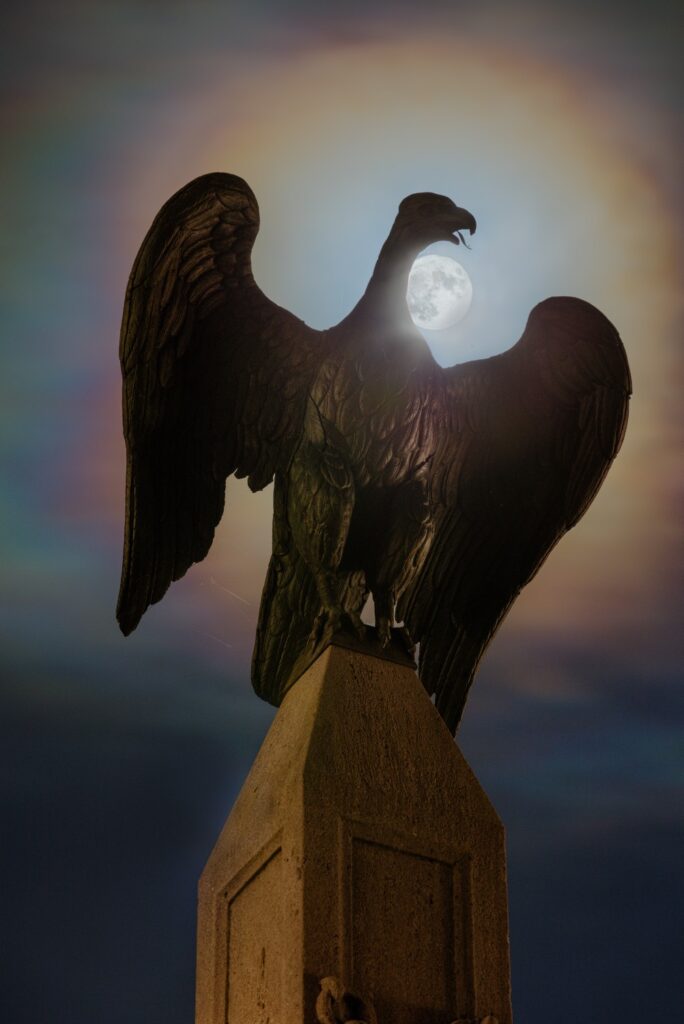 Adler-Statue auf einem Steinpfeiler bei Nacht, Mond zwischen Schnabel und Flügeln, farbiger Halo am Himmel.