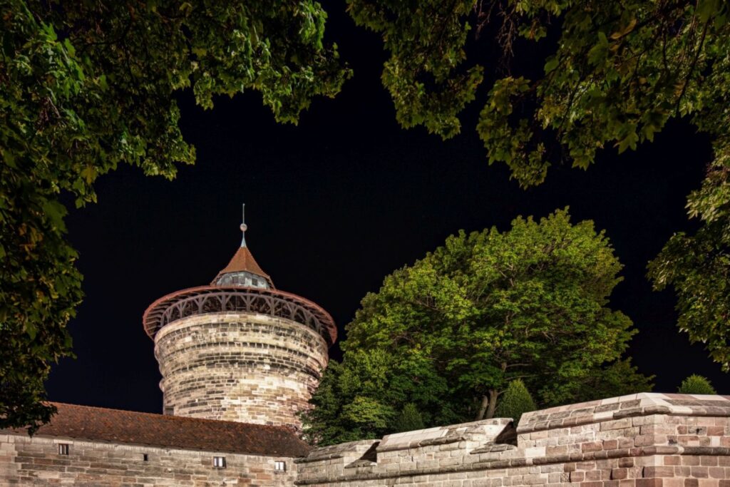 Ein alter runder Steinturm mit einem roten Kegeldach ist bei dunklem Nachthimmel sichtbar, umgeben von grünen Bäumen und einer niedrigeren Steinmauer.