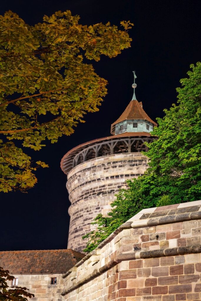 Nachts ist ein runder Steinturm mit ziegelgedecktem Spitzdach und offener Holzgalerie von Bäumen und einer alten Steinmauer umrahmt.