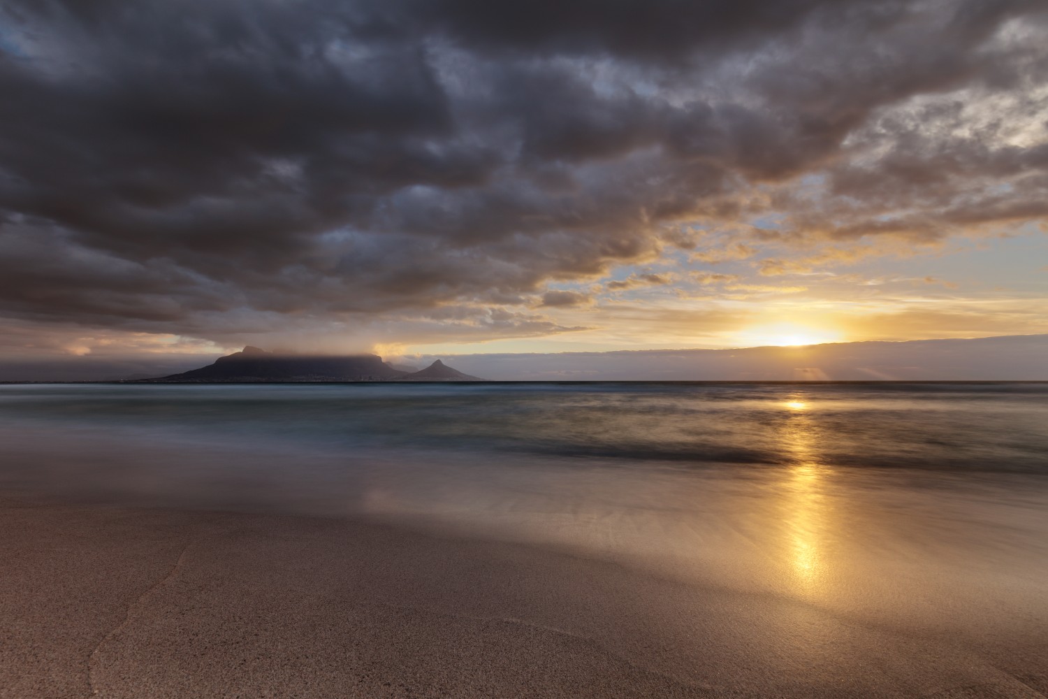 Sonnenuntergang am Strand mit goldener Wasserreflexion, dunklen Wolken und einer Insel am Horizont.
