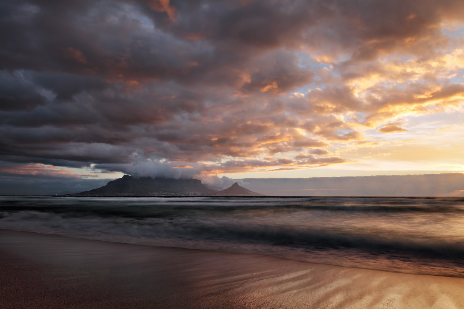 Sonnenuntergang am Strand: dunkle Wolken links, goldene Lichtstreifen rechts, sanfte Wellen und Berge am Horizont.