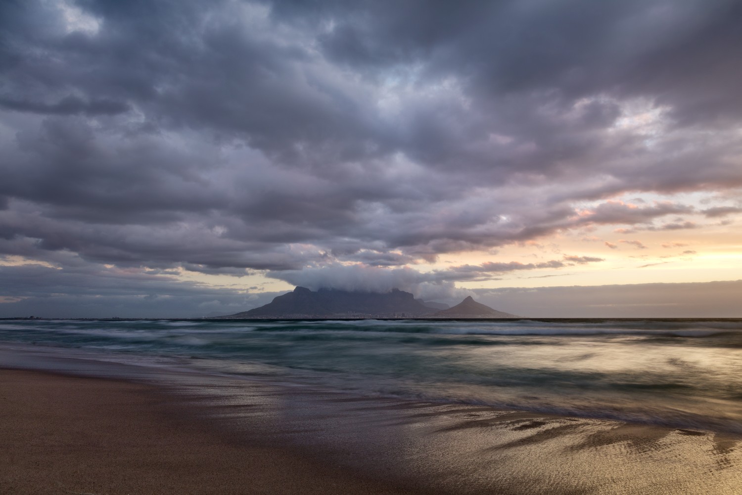 Küstenstrand bei Abendlicht: dunkle Wolken, sanfte Wellen und entfernte Bergkette am Horizont.