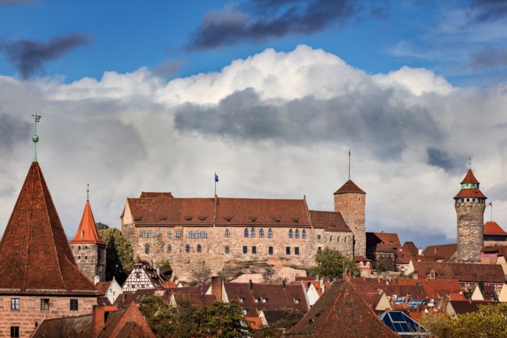 Eine imposante Burg thront über einer historischen Stadt mit zahlreichen roten Dächern unter einem weiten Himmel mit großen Wolken und blauem Himmelsabschnitten.