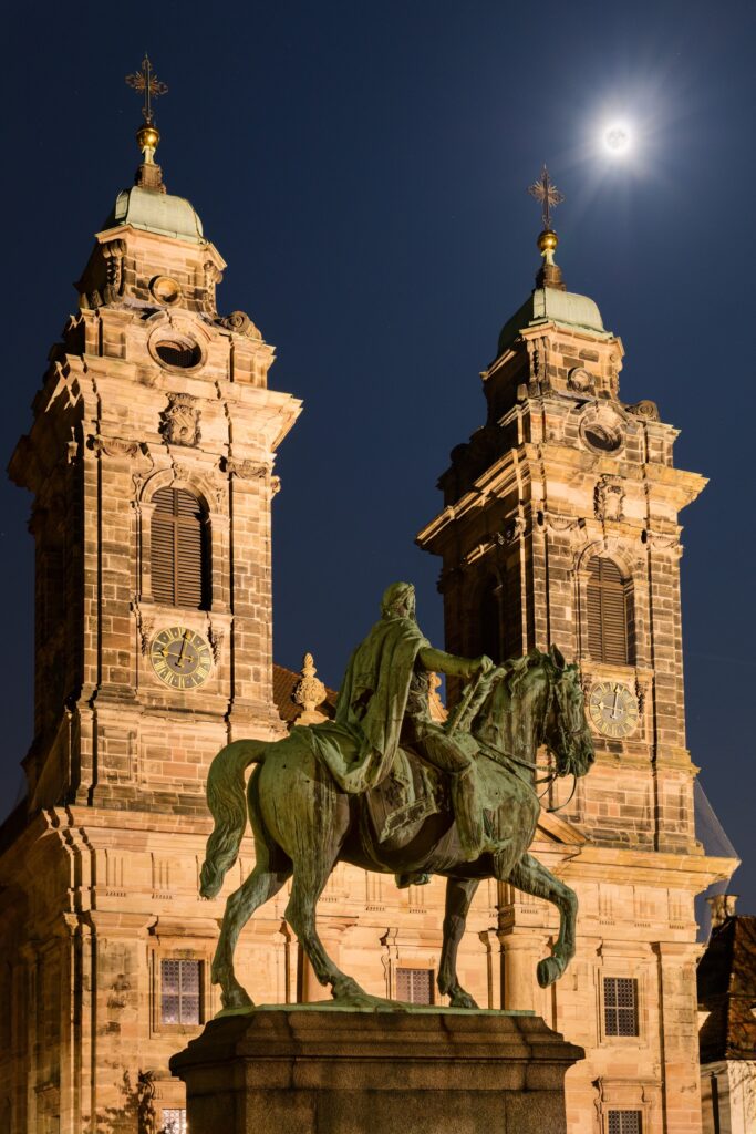 Nachtaufnahme einer barocken Kirche mit zwei Türmen, Reiterstatue im Vordergrund, Mond am Himmel, warm beleuchtet.