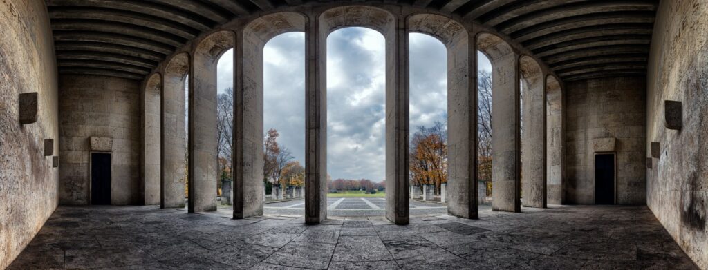 Historische Säulenarkade: Innenraum mit hohen Steinbögen, Blick auf einen herbstlichen Park mit Bäumen und Wolken.