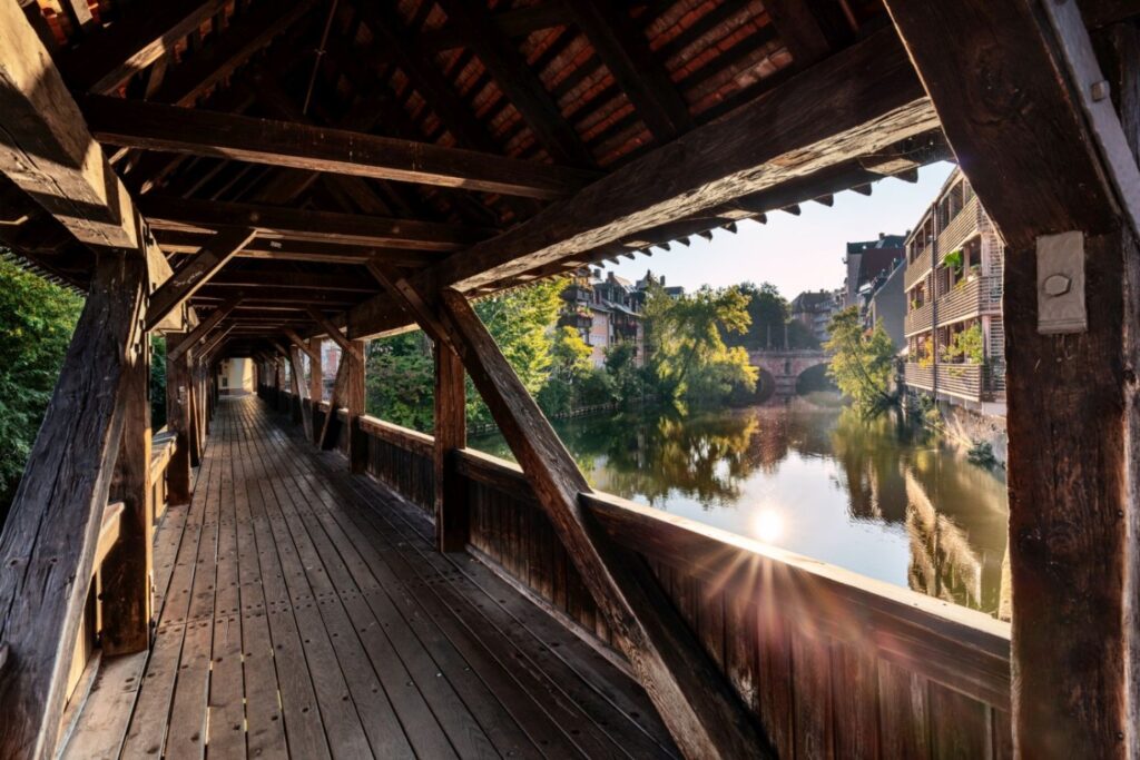 Eine lange überdachte Holzbrücke bietet einen Blick auf einen Fluss, dessen Ufer von Bäumen und Häusern gesäumt sind, sowie auf eine weitere Brücke in der Ferne, während die Sonne das Wasser zum Glänzen bringt.