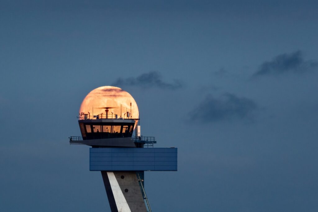 Ein großer, orangeroter Vollmond scheint hinter der Kanzel eines Fluglotsenturms vor einem dunklen, leicht bewölkten Abendhimmel.