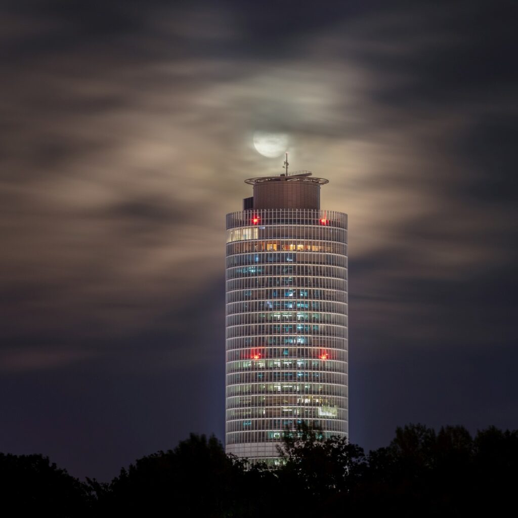 Ein hohes, zylindrisches Gebäude mit hell erleuchteten Fenstern ragt nachts unter einen von fließenden Wolken bedeckten Himmel, durch den der Mond scheint, während dunkle Baumwipfel den unteren Bildrand säumen.