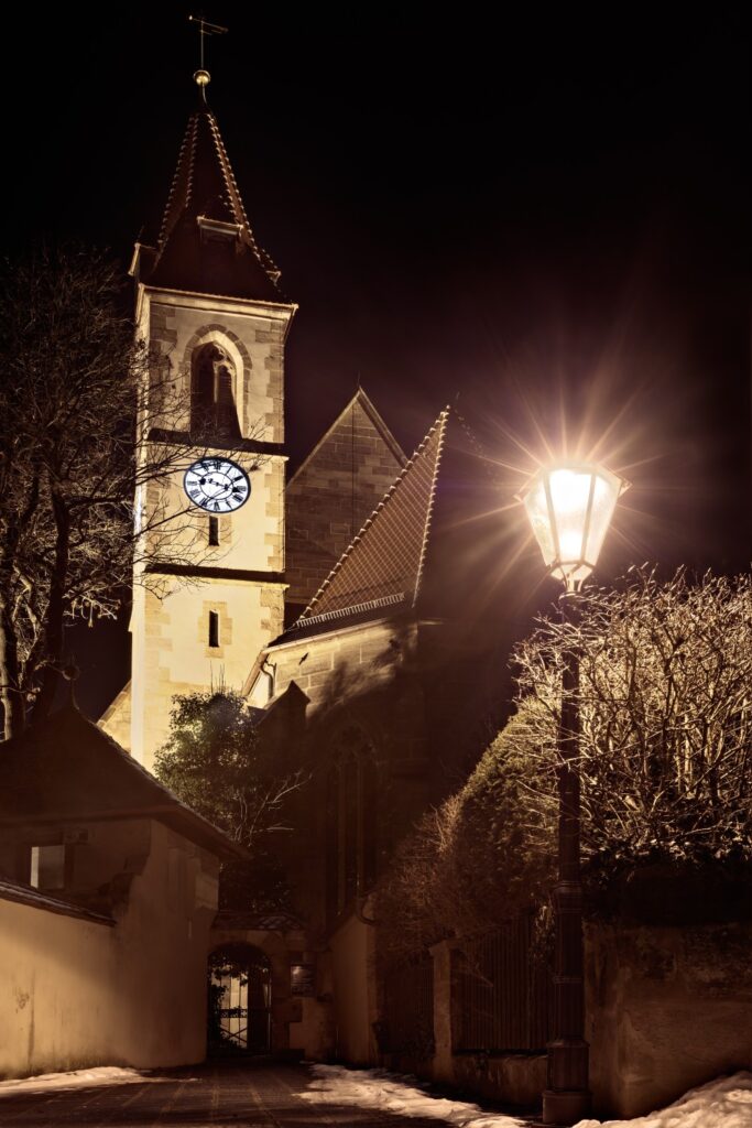 Nachtaufnahme einer historischen Kirche mit Uhrturm, beleuchteter Straßenlaterne und verschneitem Gehweg.