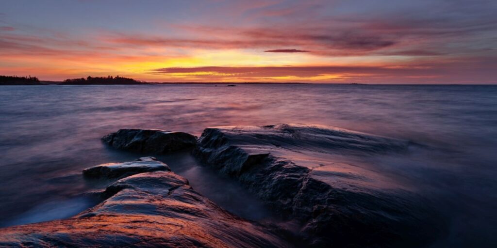 Sonnenuntergang am Meer über dunklen Felsen im Vordergrund, ruhiges Wasser und warme Farbtöne.