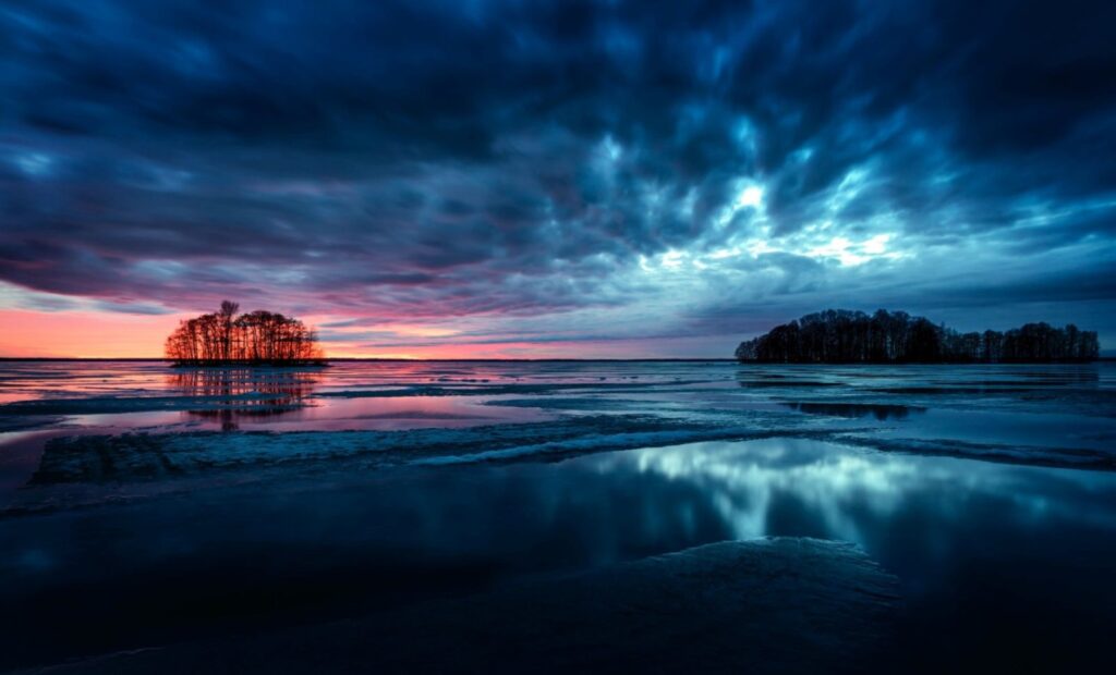 Abenddämmerung am ruhigen See mit zwei Inseln; dramatische Wolken, roter Horizont und reflektierendes Wasser.